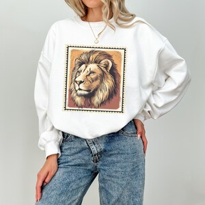 K&ouml;nnte beinhalten: Wei&szlig;er Pullover mit einem Aufdruck eines L&ouml;wenkopfes in einem Vintage-Briefmarken-Design. Der L&ouml;we ist braun und hat eine wei&szlig;e M&auml;hne.