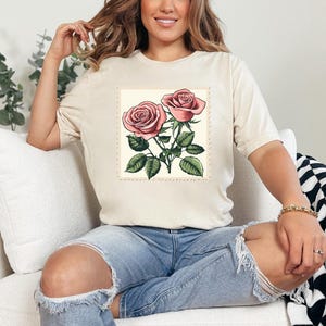 K&ouml;nnte beinhalten: Ein wei&szlig;es T-Shirt mit einer Vintage-Illustration von zwei rosa Rosen mit gr&uuml;nen Bl&auml;ttern und Stielen. Die Rosen sind von einem dekorativen Rand umgeben.