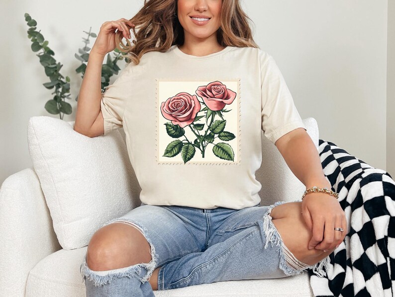 K&ouml;nnte beinhalten: Ein wei&szlig;es T-Shirt mit einer Vintage-Illustration von zwei rosa Rosen mit gr&uuml;nen Bl&auml;ttern und Stielen. Die Rosen sind von einem dekorativen Rand umgeben.