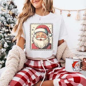 May include: White t-shirt featuring a Santa Claus graphic in a postage stamp design. The Santa illustration has a red hat and beard. The person is wearing red and white plaid pajama pants and holding a white mug with a Santa Claus graphic.