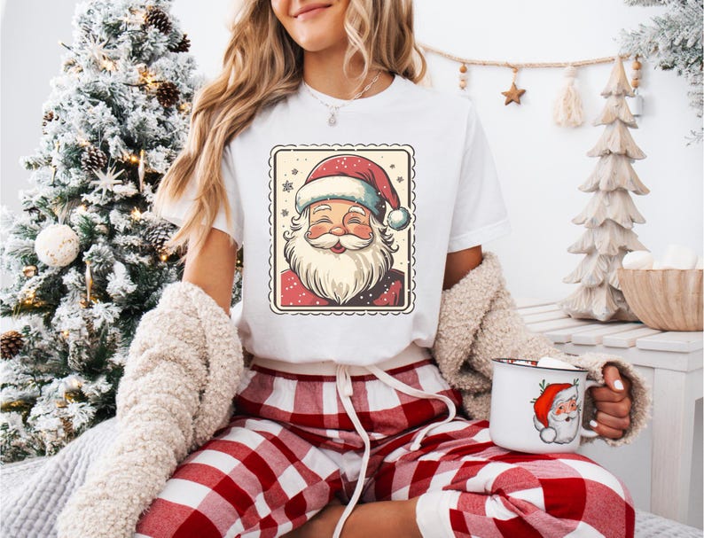 May include: White t-shirt featuring a Santa Claus graphic in a postage stamp design. The Santa illustration has a red hat and beard. The person is wearing red and white plaid pajama pants and holding a white mug with a Santa Claus graphic.