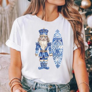 May include: White t-shirt featuring a blue and white watercolour illustration of a nutcracker holding a surfboard. The surfboard is decorated with a blue and white floral pattern.