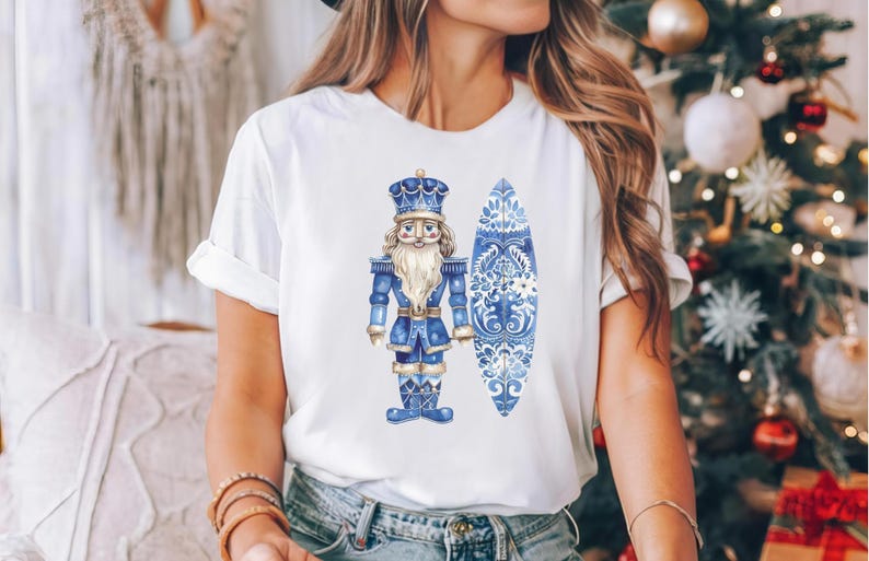 May include: White t-shirt featuring a blue and white watercolour illustration of a nutcracker holding a surfboard. The surfboard is decorated with a blue and white floral pattern.