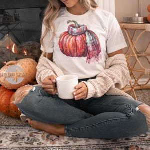 May include: White t-shirt featuring a watercolor illustration of a pumpkin wearing a plaid scarf. The pumpkin is a vibrant mix of red, orange, and pink hues. The shirt is paired with blue jeans and a cream-colored cardigan.