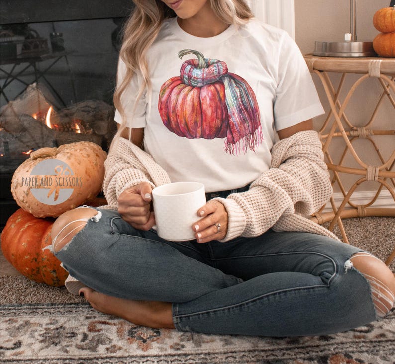 May include: White t-shirt featuring a watercolor illustration of a pumpkin wearing a plaid scarf. The pumpkin is a vibrant mix of red, orange, and pink hues. The shirt is paired with blue jeans and a cream-colored cardigan.