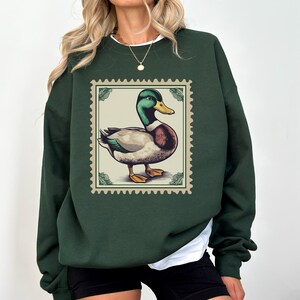 K&ouml;nnte beinhalten: Ein dunkelgr&uuml;ner Pullover mit einer Vintage-Illustration eines Enten auf einer Briefmarke. Die Ente ist braun und wei&szlig; mit einem gr&uuml;nen Kopf.
