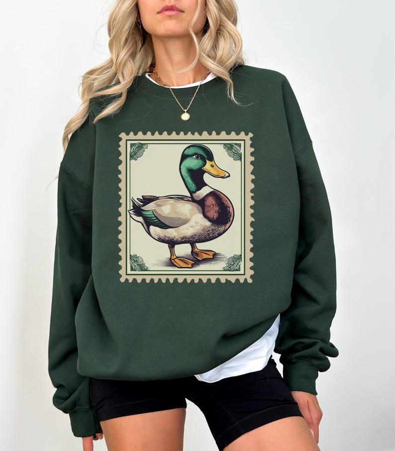 K&ouml;nnte beinhalten: Ein dunkelgr&uuml;ner Pullover mit einer Vintage-Illustration eines Enten auf einer Briefmarke. Die Ente ist braun und wei&szlig; mit einem gr&uuml;nen Kopf.
