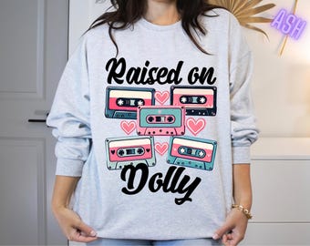 May include: A light gray sweatshirt with the text "Raised on Dolly" in black. The text is surrounded by a design of colorful cassette tapes and pink hearts.