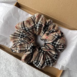 May include: A beige, black, and red plaid scrunchie sits inside a brown cardboard box lined with white tissue paper. The scrunchie is made of a soft fabric and has a gathered design.