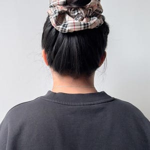 Pode incluir: Uma scrunchie xadrez bege est&aacute; em cima de um coque de cabelo escuro. A scrunchie apresenta um padr&atilde;o de xadrez bege, vermelho e preto. A pessoa est&aacute; vestindo um moletom cinza escuro.