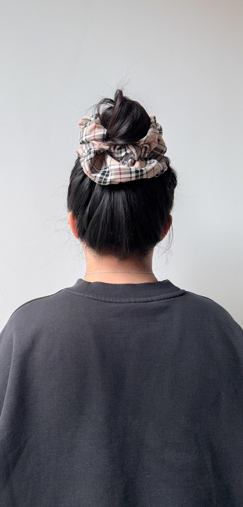 Pode incluir: Uma scrunchie xadrez bege est&aacute; em cima de um coque de cabelo escuro. A scrunchie apresenta um padr&atilde;o de xadrez bege, vermelho e preto. A pessoa est&aacute; vestindo um moletom cinza escuro.