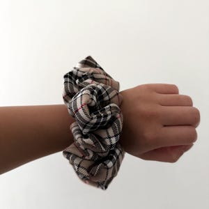 Pode incluir: Uma scrunchie xadrez bege, preta e vermelha. O tecido &eacute; franzido e enrolado em volta de um pulso. A scrunchie &eacute; um acess&oacute;rio de cabelo, e o padr&atilde;o xadrez &eacute; um design cl&aacute;ssico.
