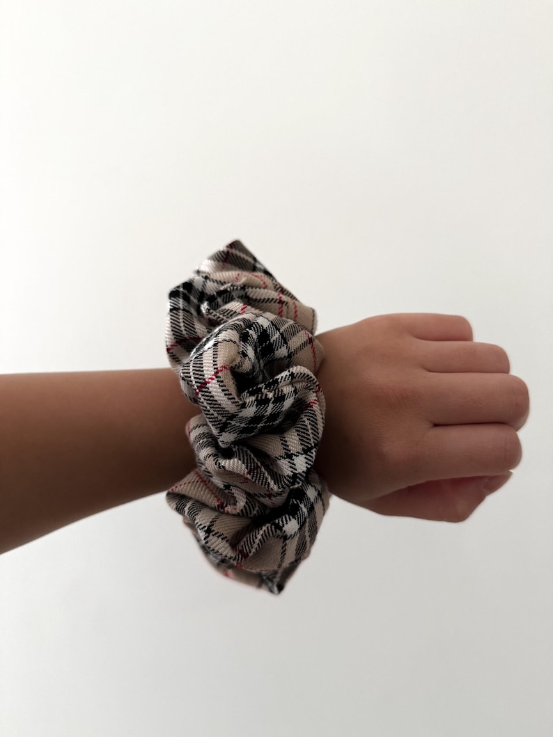 Pode incluir: Uma scrunchie xadrez bege, preta e vermelha. O tecido &eacute; franzido e enrolado em volta de um pulso. A scrunchie &eacute; um acess&oacute;rio de cabelo, e o padr&atilde;o xadrez &eacute; um design cl&aacute;ssico.