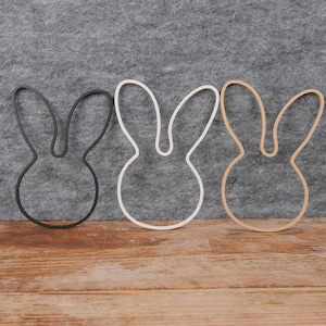 Op de afbeelding: Drie konijnvormige uitsparingen in zwart, wit en natuurlijke houttinten. De konijn silhouetten worden weergegeven op een houten oppervlak tegen een grijze vilt achtergrond. Deze decoratieve uitsparingen zijn ideaal voor Pasen of voorjaarsknutselwerk.