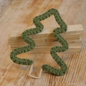 Può includere: Un segnalibro verde all'uncinetto a forma di albero stilizzato. Il segnalibro è realizzato con filato spesso e strutturato e ha un piccolo pezzo di metallo rettangolare nella parte inferiore. Il segnalibro è appoggiato su una pila di blocchi di legno.