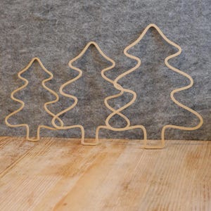 Puede incluir: Tres contornos de árboles de Navidad de madera de diferentes tamaños. Los árboles son de color marrón claro y tienen un diseño ondulado. Están dispuestos sobre una superficie de madera con un fondo gris.