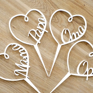 Könnte beinhalten: Vier weiße herzförmige Tortendekorationen mit den Namen "Papa", "Mama", "Oma" und "Opa" in Kursivschrift.