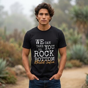May include: A black short-sleeved t-shirt featuring white text that reads WE CAN TAKE YOU TO ROCK BOTTOM and the orange text Khirbet Safra. The shirt has a crew neckline and is made of a solid black fabric with a standard fit and straight hem.