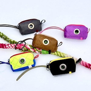 May include: Five different coloured dog poo bag holders with a round opening and a drawstring. The holders are made of different materials, including fabric and leather. The holders are on a white surface with a rope.