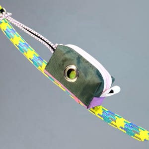 Puede incluir: Una pequeña bolsa verde oliva con cremallera rosa y ojal metálico, sujeta a una cuerda estampada amarilla y azul. La bolsa tiene un acento morado y una lengüeta blanca. La cuerda está colocada en diagonal sobre un fondo gris.