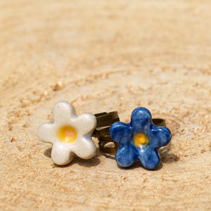 May include: Two flower-shaped rings. One ring is white with a yellow center, and the other is blue with a yellow center. The rings are set on a bronze-colored band. The background is a light brown, textured surface.