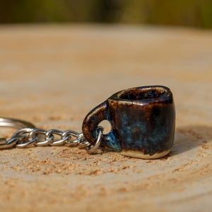 May include: A miniature ceramic coffee cup keychain with a dark brown and blue glaze. The small mug has a handle and is attached to a silver chain and keyring. The keychain is resting on a light brown wooden surface.