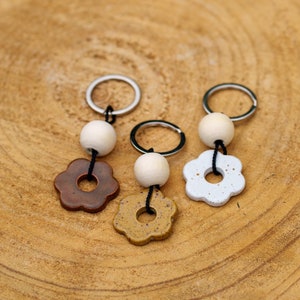 May include: Three keychains with white ceramic beads and flower-shaped charms. The charms are brown, white with black speckles, and yellow with black speckles.