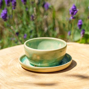 Può includere: Un piccolo set fatto a mano di ciotola e piattino in ceramica. La ciotola e il piattino sono smaltati in tonalità di verde e giallo, con un effetto craquelé. Il set è esposto su una superficie di legno, con fiori viola sullo sfondo.