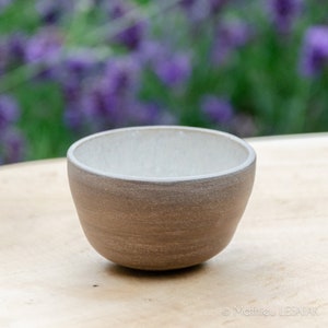 Peut inclure: Un petit bol en céramique brun clair avec un intérieur blanc. Le bol est posé sur une surface en bois avec un arrière-plan flou de fleurs violettes.