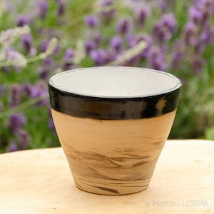 May include: A small, white ceramic cup with a black rim and brown marbling. The cup is sitting on a wooden surface.