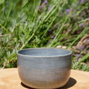 Peut inclure: Un bol en céramique fait à la main avec un glaçage bleu-gris mat. Le bol a une forme arrondie et repose sur une surface en bois, avec un arrière-plan flou de feuillage vert et de fleurs violettes.