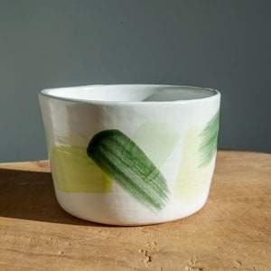 Peut inclure: Un bol en céramique blanche avec un motif abstrait vert et jaune. Le bol est posé sur une surface en bois.