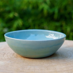 May include: A light blue ceramic bowl with a subtle glaze variation. The bowl is sitting on a wooden surface.