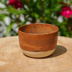 Puede incluir: Un cuenco de cerámica hecho a mano con un diseño de dos tonos. La parte superior es de un marrón intenso, mientras que la sección inferior es de un blanco roto cremoso. El cuenco está sobre una superficie de madera, con un fondo borroso verde y rosa.