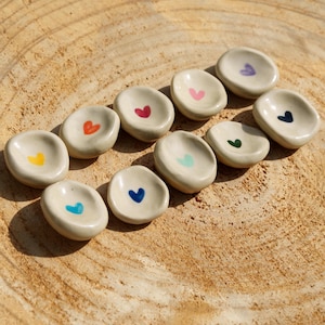 Peut inclure: Une collection de dix petits plats en céramique blanc cassé de forme ovale, chacun orné d'un cœur peint à la main dans une couleur différente, notamment jaune, orange, rouge, rose, bleu clair, bleu foncé, vert menthe, vert foncé, violet et noir.