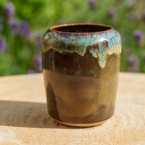 May include: A cylindrical ceramic cup featuring a glossy finish with a deep brown base color that transitions into a vibrant turquoise and cream drip glaze pattern around the upper rim, creating a smooth, reflective surface on the exterior.