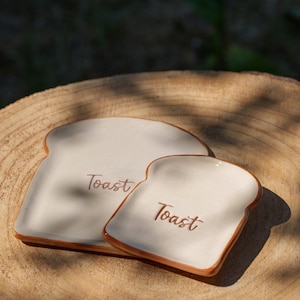 Puede incluir: Dos platos de cerámica con forma de tostada. Los platos son de color blanquecino con ribete marrón y la palabra "Toast" en escritura marrón. Los platos están sobre una superficie de madera.