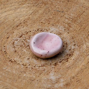 Peut inclure: Un petit plat rond en céramique rose avec un bord légèrement surélevé. Le plat a une texture irrégulière et mouchetée et repose sur une surface en bois.