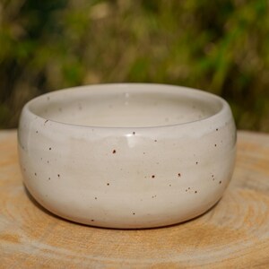 May include: A small, off-white ceramic bowl with a speckled design. The bowl has a rounded shape and sits on a wooden surface. The speckled pattern features small, dark brown spots.