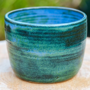 Könnte beinhalten: Eine handgefertigte Keramiktasse mit einer lebendigen, wirbelnden Glasur in Blau- und Grüntönen. Die Tasse hat eine abgerundete Form und eine glatte, glänzende Oberfläche. Das Innere ist leuchtend blau und kontrastiert mit dem dunkleren Äußeren. Die Tasse steht auf einer Holzoberfläche.