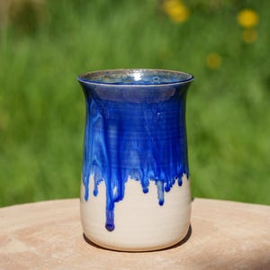 Peut inclure: Un vase en céramique blanche avec un glaçage bleu qui coule sur les côtés. Le vase a un fond arrondi et une large ouverture.