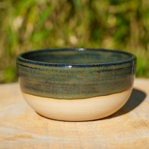 Könnte beinhalten: Eine handgefertigte Keramikschale mit zweifarbigem Design. Der obere Teil ist in einem melierten Türkis gehalten, während die untere Hälfte cremefarben ist. Die Schale ist rund und steht auf einer Holzoberfläche, mit einem verschwommenen grünen Hintergrund.