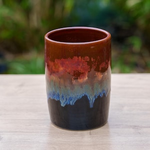 Peut inclure: Un vase en céramique avec un glaçage rouge, bleu et noir. Le vase a une large ouverture et une forme cylindrique.
