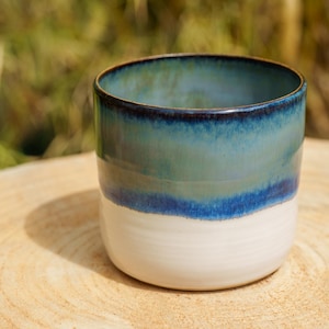 Puede incluir: Una taza cilíndrica de cerámica con un esmalte de dos tonos. La parte superior muestra un degradado de azul marino profundo y verde azulado, mientras que la parte inferior es de un color blanco roto sólido y liso. El borde es oscuro.