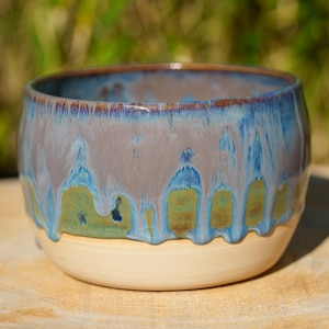 Peut inclure: Un bol en céramique fait à la main de forme arrondie. Le bol présente une base crème, une section intermédiaire gris-bleu avec des gouttes bleues et vertes, et un bord bleu foncé. Le bol est posé sur une surface en bois.