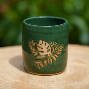 May include: A green ceramic mug with a gold leaf design. The mug is sitting on a wooden surface.