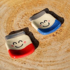 Peut inclure: Deux petits vide-poches en céramique en forme de pot avec des visages souriants et des joues roses. Un pot a une base rouge, l'autre bleue. Les deux ont des couvercles gris et reposent sur une surface texturée brun clair.