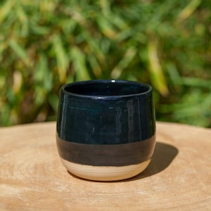 Peut inclure: Un gobelet en céramique fait à la main, avec une partie supérieure bleu foncé, une bande noire au milieu et une base beige naturel. Le gobelet est posé sur une surface en bois, avec un arrière-plan vert flou.