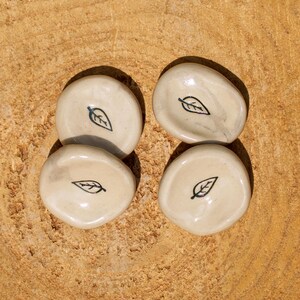 Puede incluir: Cuatro pequeños cuencos de cerámica blanquecina con un diseño de hoja negra. Están dispuestos sobre una superficie de madera marrón claro texturizada. El diseño de la hoja es simple y elegante.