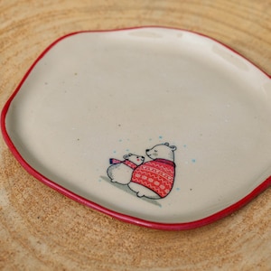 May include: A small, off-white ceramic plate with an irregular, rounded shape and a thin, hand-painted red rim. The center of the plate features a detailed illustration of two polar bears, one wearing a red patterned sweater and one a scarf.